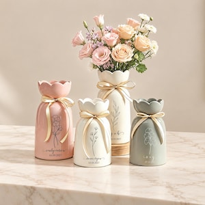 Puede incluir: Cuatro jarrones de cerámica con bordes festoneados y lazos de cinta dorada. Uno es rosa, otro gris y dos son de color crema. Cada jarrón presenta una delicada ilustración floral de líneas, nombres personalizados y una fecha en cursiva.