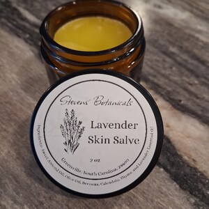 May include: An amber glass jar containing yellow skin salve with a black lid. The label reads Stevens' Botanicals, Lavender Skin Salve, 2 oz, Greenville, South Carolina, 29690, and lists ingredients including Sweet Almond Oil, Olive Oil, Beeswax, Calendula, Thyme, and Lavender Essential Oil.