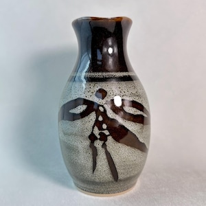 May include: A ceramic vase with a glossy dark brown neck and a speckled light grey body. The body features a dark brown, abstract, symmetrical design resembling a dragonfly or floral motif, with a smooth, rounded shape and a flared rim.