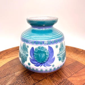 May include: A ceramic vase with a rounded body and a flared rim, featuring a white base decorated with teal and blue floral patterns, horizontal bands, and a repeating circular motif around the upper and lower sections of the vessel.