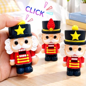 Peut inclure: Trois petites figurines de casse-noisette avec des vestes rouges à bordures jaunes, des chapeaux noirs avec des étoiles jaunes ou des plumes rouges, et des cheveux blancs. Chaque figurine a une moustache blanche et une bouche rectangulaire.