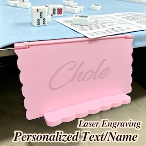 May include: A light pink plastic mahjong tile rack with a scalloped edge design, featuring the name Chole engraved in a cursive script on the front surface. The rack is designed to clip onto the edge of a table, holding game tiles upright.