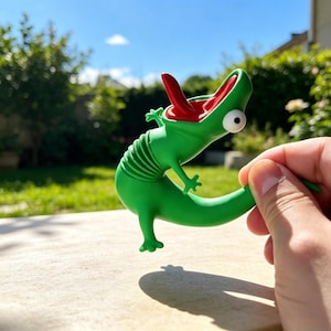 May include: A green lizard-shaped toy with a flexible, accordion-style midsection, a wide-open mouth revealing a red interior and tongue, and a single large white eye. The figure has four small limbs and is held in a horizontal position.