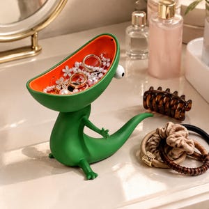May include: A green dinosaur-shaped trinket dish with an orange interior, featuring a single white eye with a black pupil. The dish holds various silver-toned rings and necklaces, and the figure has a long tail and small arms.