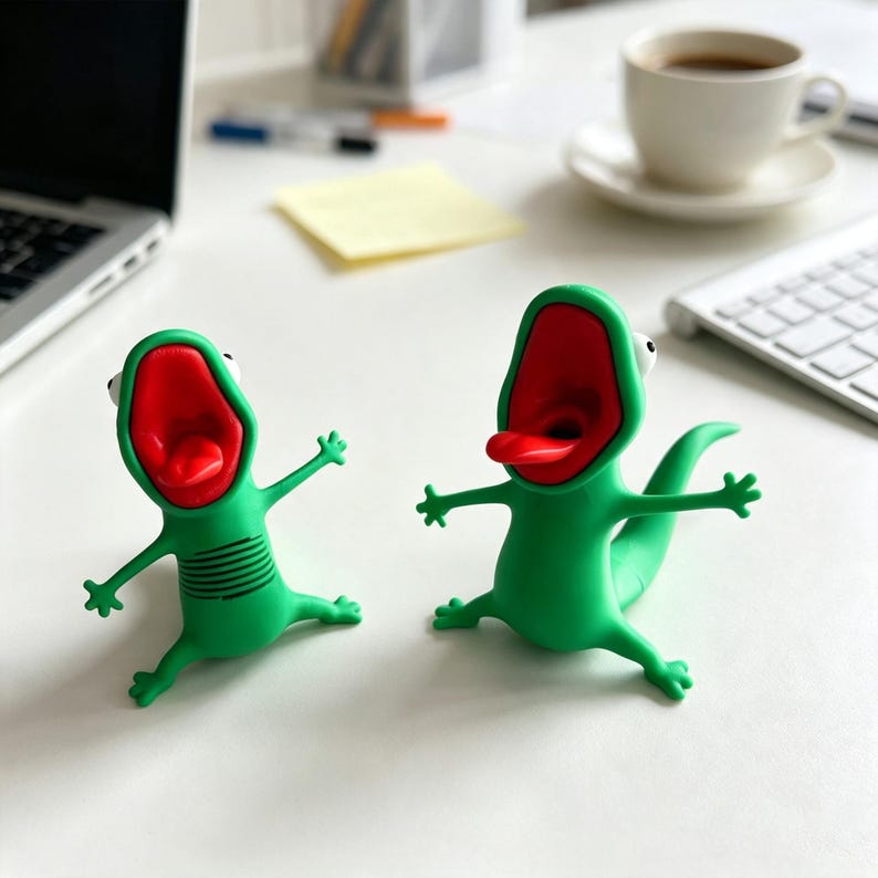 May include: Two green lizard-shaped figures with wide-open mouths, bright red interiors, and protruding tongues. Each figure has small arms and legs extended outward, white eyes, and a smooth finish. One figure features horizontal lines on its torso.