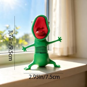 May include: A green frog-like figurine with a large, open red mouth and a protruding tongue. The figure has a ribbed midsection and outstretched arms. It measures 3.74 inches in height and 2.95 inches in width as indicated by the text.