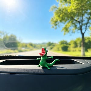 May include: A small green lizard figurine with a wide-open red mouth, a protruding tongue, and one large white eye. The creature has a segmented body, four short limbs, and a curved tail, positioned in a standing pose on a dark dashboard surface.