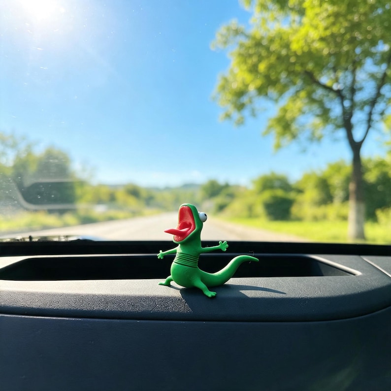 May include: A small green lizard figurine with a wide-open red mouth, a protruding tongue, and one large white eye. The creature has a segmented body, four short limbs, and a curved tail, positioned in a standing pose on a dark dashboard surface.