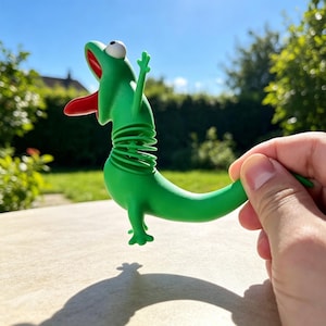 May include: A green lizard-shaped toy with a flexible coiled spring midsection, a wide-open mouth featuring a red tongue, and a single visible white eye. The toy has a smooth matte finish, small limbs, and a long tail, designed with a spring mechanism.