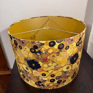May include: A cylindrical drum lampshade featuring a repeating pattern of gold, yellow, and black circular motifs, swirls, and dark floral shapes. The interior of the shade is a solid mustard yellow, supported by a gold-toned metal spider fitter.