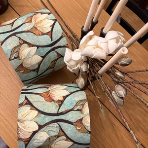 May include: Two cylindrical lampshades featuring a repeating pattern of white magnolia flowers with dark brown branches against a light blue background, positioned next to a candelabra with white taper candles and decorative floral accents.