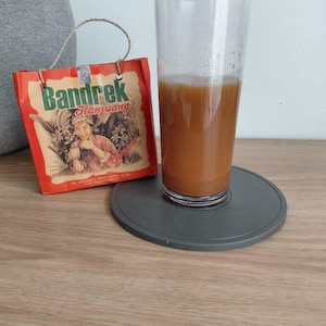 May include: A tall clear glass filled with a light brown beverage sits on a circular gray coaster next to a small orange paper bag with a string handle. The bag features the text Bandrek Hanjuang and an illustration of a person holding a cup.