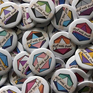 May include: A collection of round pin-back buttons featuring a multi-faceted 20-sided die graphic in various color combinations representing pride flags. Each button displays a banner across the center with the text Roll with Pride in black.