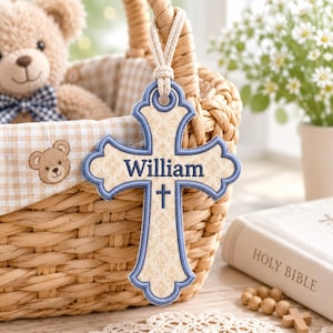 May include: A decorative cross ornament featuring a cream-colored fabric with a subtle gold fleur-de-lis pattern, bordered by blue stitching. The name William is embroidered in blue thread above a small blue cross symbol. It hangs from a thick rope.