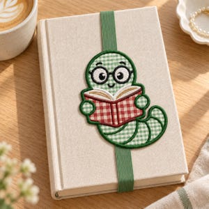 May include: A beige fabric-covered notebook featuring an embroidered green and white gingham worm wearing black glasses and reading a red and white gingham book. A vertical green elastic band secures the notebook, centered over the design.