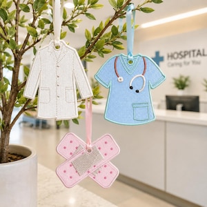 May include: Three hanging ornaments shaped like a white lab coat, a light blue scrub top with a stethoscope, and a pink adhesive bandage with polka dots. Each ornament features stitched details and is suspended by a matching satin ribbon loop.