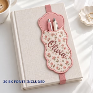 Puede incluir: Un cuaderno cubierto de tela beige con un portalápices festoneado de borde rosa y estampado floral. El nombre Olivia está bordado en letra marrón sobre el soporte, que contiene dos bolígrafos. El texto 30 BX FONTS INCLUDED es visible.
