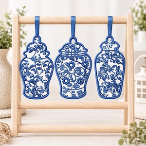 May include: Three blue embroidered ornaments shaped like ginger jars, featuring intricate floral and vine patterns. Each ornament is suspended by a matching blue satin ribbon from a light-colored wooden display stand with horizontal bars.