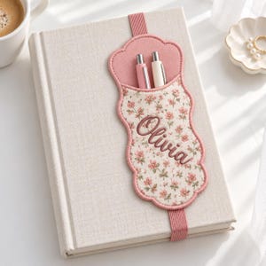 May include: A beige fabric-covered notebook featuring an attached elastic band with a scalloped, floral-patterned fabric pocket. The pocket is embroidered with the name Olivia in brown script and holds two pens, one pink and one white.