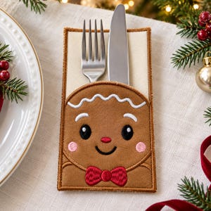 May include: A rectangular fabric cutlery holder featuring an embroidered gingerbread person design. The brown gingerbread character has a white icing-style trim, black eyes, pink cheeks, a red nose, and a red bow tie. A silver fork and knife are tucked inside.