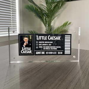 May include: A clear acrylic display block featuring a replica movie ticket for the 1931 film Little Caesar. The ticket includes a portrait of Edward G. Robinson, the title, director Mervyn LeRoy, venue details, a barcode, and a price of $0.50.
