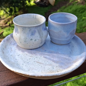 May include: A ceramic set consisting of a round plate and two small cups. The plate and the left cup feature a white base with blue speckled glaze and drips. The right cup has a smooth, light blue glaze with a subtle horizontal ridge.