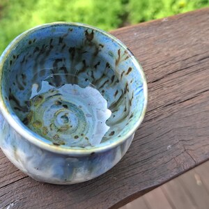May include: A small, round ceramic bowl featuring a variegated glaze in shades of light blue, white, and dark brown. The interior displays a swirling pattern with dark brown speckles and streaks against a light blue base, with a smooth rim.