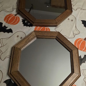 May include: Two octagonal mirrors featuring medium-toned wooden frames with a visible grain. The mirrors have a smooth, reflective surface and are shaped with eight equal sides, providing a geometric aesthetic for wall mounting.