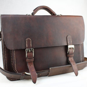 May include: A dark brown leather briefcase with a flap closure, two buckle straps, and a shoulder strap. The bag has a top handle and white stitching details. The leather has a distressed appearance.