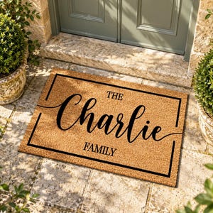 May include: A rectangular tan coir doormat featuring a black rectangular border. The text "THE" is printed in small black capital letters above the large black script name "Charlie" and the word "FAMILY" in black capital letters below.