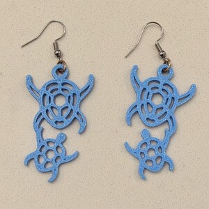 Puede incluir: Un par de pendientes colgantes de color azul claro con un diseño recortado de una tortuga marina grande con una tortuga más pequeña unida debajo. Los pendientes cuelgan de ganchos de metal plateado y tienen un acabado mate texturizado.