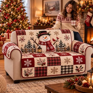 May include: A quilted sofa cover featuring a festive holiday design with a central snowman, red and green plaid squares, and white snowflake patterns. The fabric displays pine trees, holly berries, and a lantern motif on a cream-colored background.