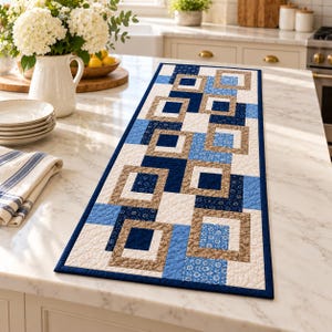 May include: A rectangular quilted table runner featuring a geometric pattern of nested squares in shades of navy blue, light blue, tan, and white. The fabric displays a mix of solid colours and small floral prints with a dark blue border trim.