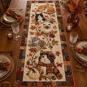 May include: A rectangular quilted table runner featuring a central cream-colored panel with an embroidered design of five cats nestled among autumn leaves, acorns, and a pumpkin. The border consists of a patchwork pattern in shades of brown and orange.