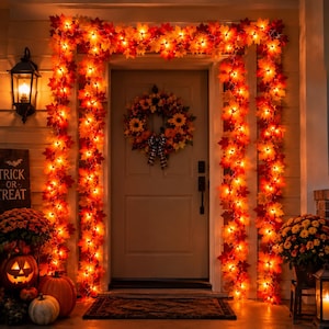 Puede incluir: Una puerta decorada con una guirnalda de hojas de arce artificiales iluminadas en tonos naranja, rojo y amarillo. En la puerta cuelga una corona con girasoles, calabazas y un lazo a cuadros blanco y negro, flanqueada por calabazas y crisantemos.