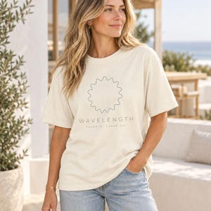 May include: A cream-colored short-sleeve t-shirt featuring a dark gray wavy circular line graphic centered on the chest. Below the graphic, the text reads "WAVELENGTH" in capital letters, followed by the smaller text "tuned in, tuned out."