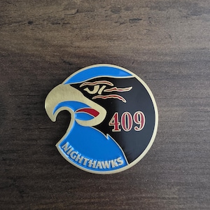 Può includere: Una spilla metallica circolare con un design stilizzato di testa di falco in blu, nero e oro. Il falco ha un becco dorato, un'area oculare nera con accenti rossi e il testo 409 in rosso. La parola NIGHTHAWKS è stampata in blu sul bordo inferiore.