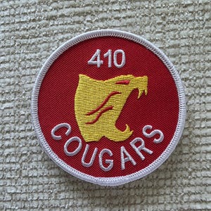May include: A circular embroidered patch with a red background and a white stitched border. The patch features a yellow cougar head silhouette in the centre, the number 410 in white above it, and the word COUGARS in white text below it.