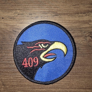 Puede incluir: Un parche bordado circular que presenta el perfil de una cabeza de águila negra sobre un fondo azul. El águila tiene un pico amarillo, detalles oculares rojos y el número 409 en rojo cosido bajo el ojo. El parche tiene un borde negro grueso.