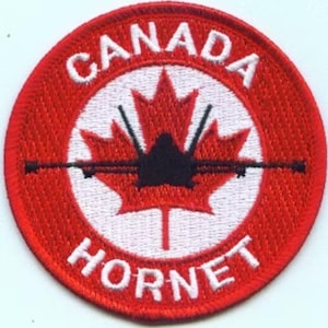 May include: A circular embroidered patch featuring a red border and a white centre. A red maple leaf is centred with a black silhouette of a fighter jet superimposed over it. The text CANADA is arched at the top and HORNET is at the bottom in white.