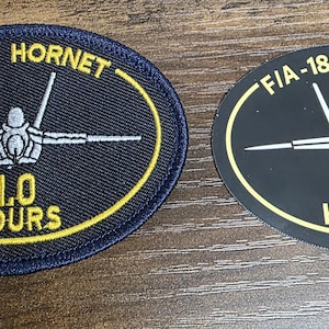 May include: Two oval-shaped patches featuring a graphic of an F/A-18 Hornet aircraft in white, with the text F/A-18 HORNET and 1.0 HOURS in yellow. One patch is embroidered with a dark blue border, while the other is a smooth, flat black material.
