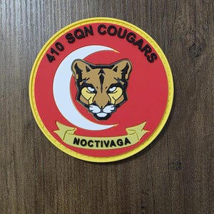 May include: A circular patch featuring a red background with a yellow border. In the centre is a tan cougar head graphic. A white crescent moon sits behind the cougar. The text 410 SQN COUGARS is arched at the top, and NOCTIVAGA is on a banner below.