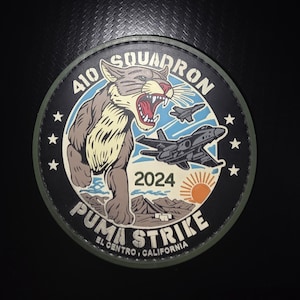 Peut inclure: Un écusson circulaire représentant un puma rugissant, deux avions de chasse et un soleil au-dessus d'un paysage désertique. Le texte indique 410 SQUADRON, 2024, PUMA STRIKE et EL CENTRO, CALIFORNIA. Le design inclut des étoiles blanches sur fond noir.