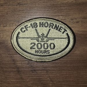 Peut inclure: Un écusson brodé de forme ovale en vert olive avec une bordure noire. L'écusson présente une silhouette noire d'un avion CF-18 Hornet au centre, avec le texte CF-18 HORNET arqué au-dessus et 2000 HOURS imprimé en noir en dessous.