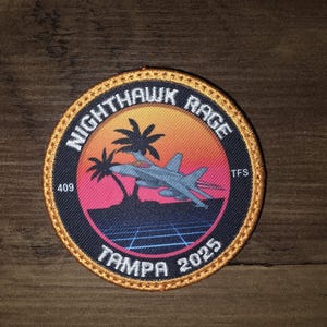 May include: A circular embroidered patch with a gold border featuring a sunset background, palm trees, and a fighter jet. The text reads NIGHTHAWK RAGE, 409, TFS, and TAMPA 2025. The design includes a dark blue grid pattern at the bottom.