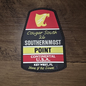 Puede incluir: Un parche en forma de escudo con un icono de cabeza de puma amarillo sobre el texto Cougar South 26 SOUTHERNMOST POINT CONTINENTAL U.S.A. KEY WEST, FL Home of the Sunset. El diseño utiliza bandas horizontales rojas, negras, blancas y amarillas.