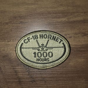 May include: An oval-shaped embroidered patch in olive green with a dark border. The patch features a central graphic of a fighter jet silhouette, with the text CF-18 HORNET arched above and the numbers 1000 followed by the word HOURS below.