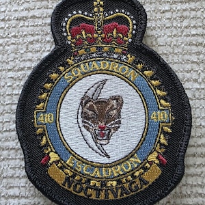 May include: A circular embroidered patch featuring a central white circle with a brown cougar head and a silver crescent moon. The border is light blue with the text SQUADRON 410 and ESCADRON 410. A gold crown sits atop the patch with the text NOCTIVAGA.