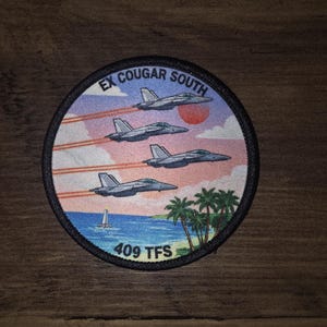 May include: A circular embroidered patch featuring four fighter jets flying in formation against a sunset sky. The text EX COUGAR SOUTH is at the top and 409 TFS is at the bottom. Below the jets is a blue ocean, a small sailboat, and palm trees.