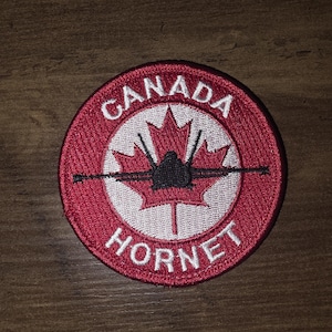 Puede incluir: Un parche bordado circular con un borde rojo que presenta el texto blanco CANADA en la parte superior y HORNET en la inferior. El centro muestra una silueta de hoja de arce blanca con un gráfico de avión de combate negro superpuesto.